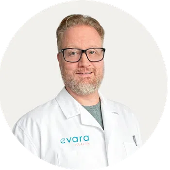 Smiling man with light red hair and beard wearing a white lab coat with Evara Health logo