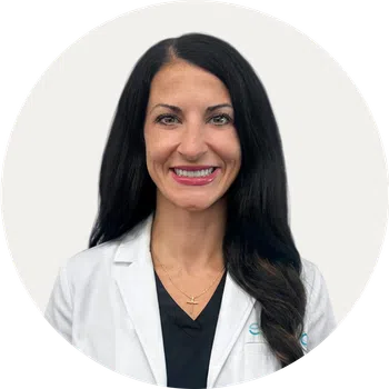 Smiling woman with long black hair wearing a white lab coat and black shirt against a plain background
