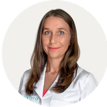 Smiling woman with long brown hair wearing a white lab coat against a plain white background.