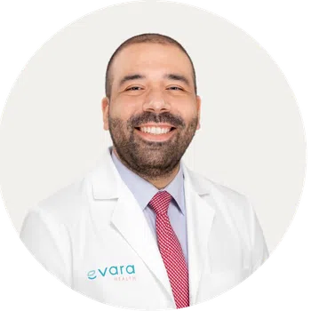 Smiling male doctor wearing white coat and red tie with Evara Health logo on coat against white background