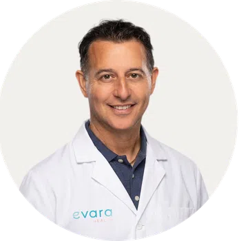 Smiling male doctor wearing a white lab coat with Evard Health logo on a plain light background.