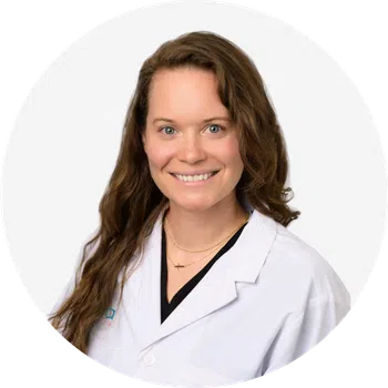 Smiling female doctor with long brown hair wearing a white lab coat against a white background.
