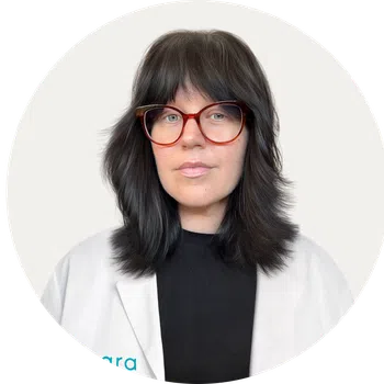Woman with dark layered hair and red glasses wearing a white coat over black top in a circular frame.