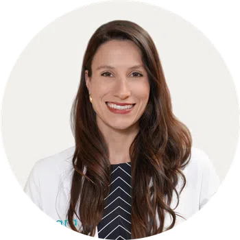 Smiling woman with long brown hair wearing a white coat and striped black shirt against a white background