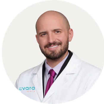 Smiling male doctor wearing a white lab coat and purple tie against a light background
