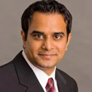 Professional headshot of a man in a dark suit with a red striped tie on a gray background.