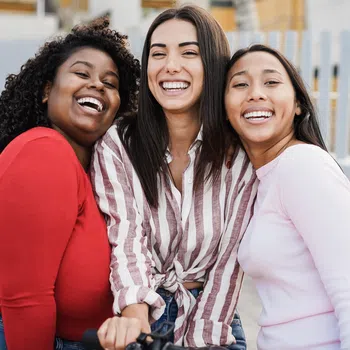 Three diverse young women smiling and hugging outdoors near a white fence, enjoying a sunny day together.