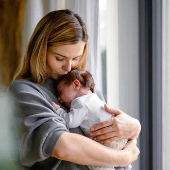 Mother tenderly holding and kissing newborn baby in cozy home by window with natural light.