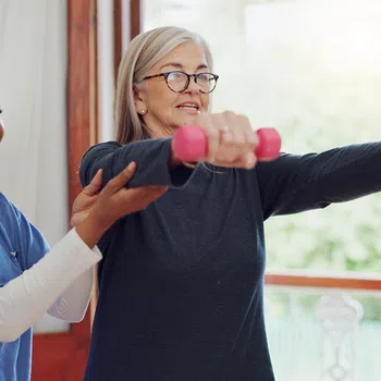 Senior woman lifting pink dumbbells guided by a smiling female fitness trainer indoors near window.