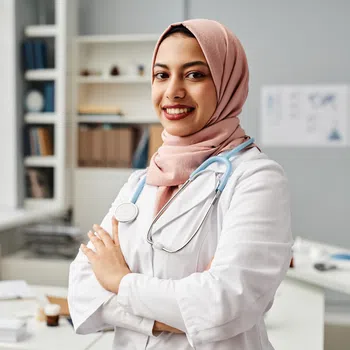 Young female doctor wearing a pink hijab and white coat with stethoscope smiling in a bright medical office.