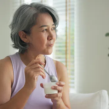 Mature woman with gray hair eating yogurt on a couch in a bright, cozy living room with plants.
