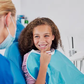 Smiling young girl sitting in dental chair wearing a blue bib while dentist in mask talks to her.