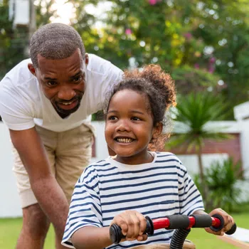 Father supports smiling daughter riding a red tricycle in a sunny backyard with greenery and flowers.