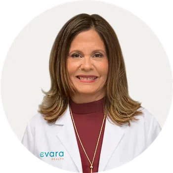 Smiling female healthcare professional wearing white coat and maroon top against a plain background.