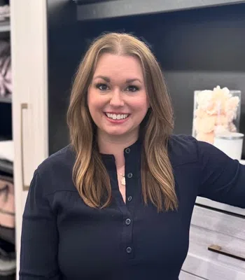 Smiling woman with long light brown hair wearing a dark blouse standing in a modern kitchen with white cabinets.