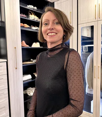 Smiling woman in sheer black polka dot top posing in a modern walk-in closet with shoes and clothes.