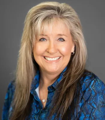 Smiling middle-aged woman with long blonde hair wearing a blue patterned blouse against a gray background