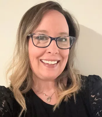 Smiling woman with glasses and wavy blonde hair wearing a black lace top and heart necklace indoors.