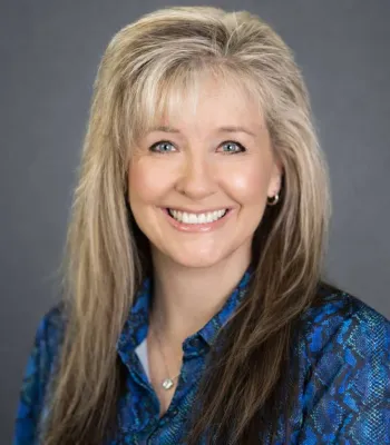 Smiling woman with long blonde hair wearing a blue patterned blouse against a plain gray background