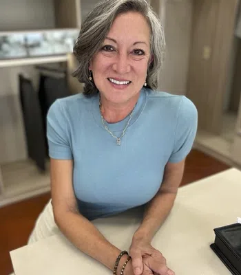 Smiling mature woman with gray hair wearing a blue shirt leaning on a table in a cozy room.