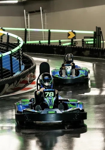 Two go-karts racing on a shiny indoor track with vibrant lighting and a curved barrier.