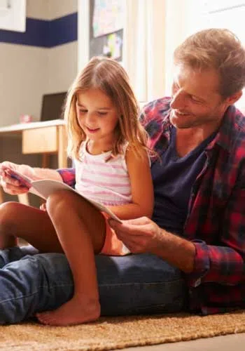 Father and daughter sitting on the floor reading a book together in a cozy room filled with natural light.