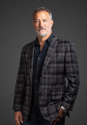 Middle-aged man with gray hair and beard wearing a plaid blazer and dark shirt, standing against a gray background.