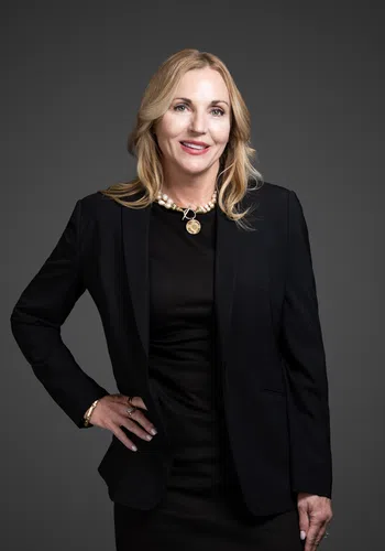 Professional woman with blonde hair wearing a black blazer and dress, pearl necklace, smiling against gray background