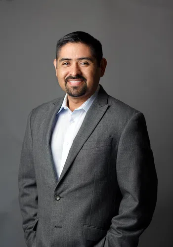 Smiling man in a gray blazer and white shirt posing against a plain gray background.