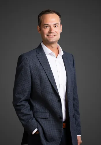 Smiling man in a dark checkered blazer and white shirt posing against a solid gray background.