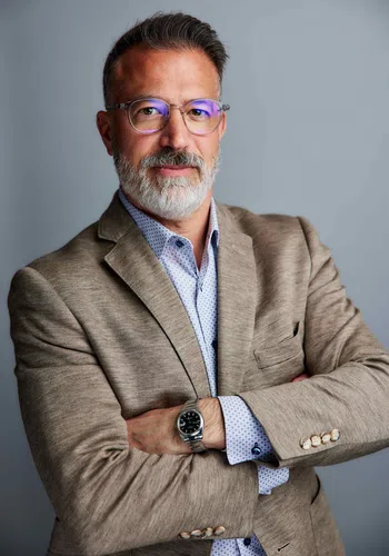 Professional mature man with glasses and gray beard wearing beige blazer and watch, arms crossed, smiling confidently