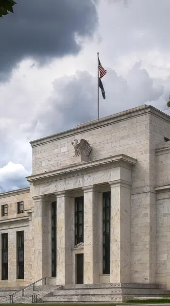 Historic Federal Reserve building with tall columns and an American flag under dramatic cloudy sky.