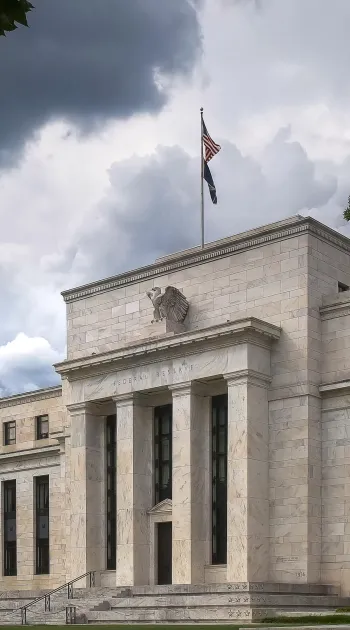 Historic Federal Reserve building with tall columns and an American flag under dramatic cloudy sky.