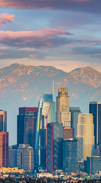 Downtown Los Angeles skyline with tall skyscrapers and mountains in the background under a colorful sky.