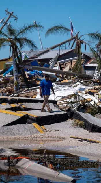 Man walking on broken road amidst hurricane destroyed houses and palm trees under blue sky