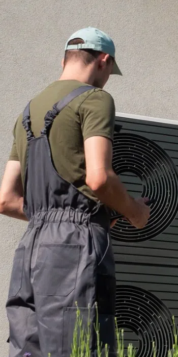 Technician in overalls inspecting outdoor air conditioning unit while homeowner watches by house wall.