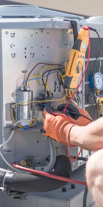 Technician repairing and testing an outdoor air conditioning unit using electrical tools and gauges.