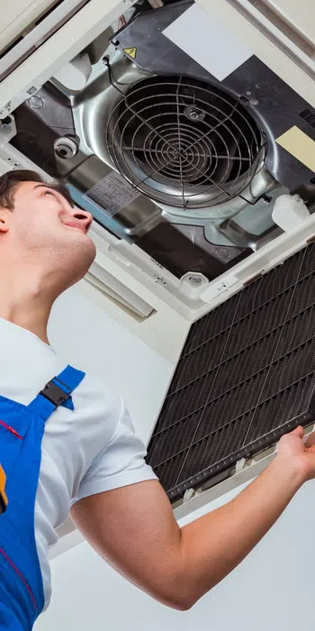 Technician in blue overalls inspecting and cleaning a ceiling air conditioning unit while holding a toolbox on a ladder.