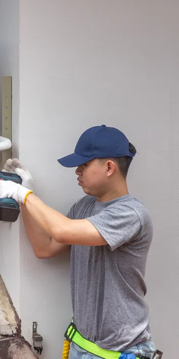Technician wearing gloves and cap installs an air conditioning unit on a house exterior wall above a tiled roof.