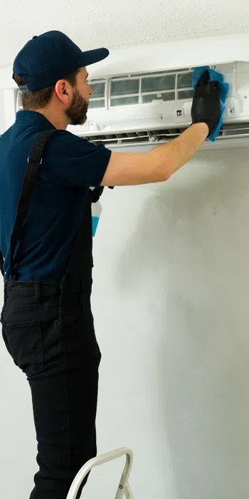 Technician in uniform servicing a wall-mounted air conditioner unit on a gray wall inside a living room.