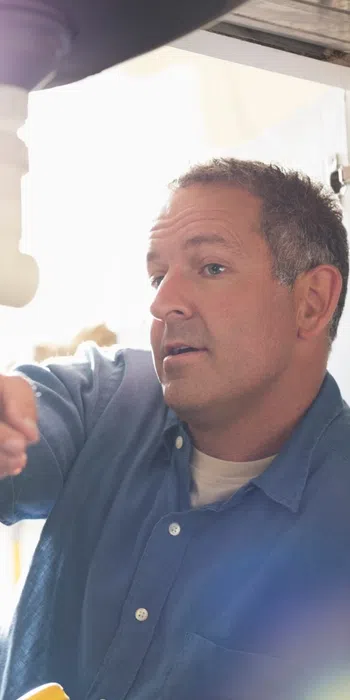 Plumber explaining repair work under a kitchen sink to a woman observing closely during maintenance.