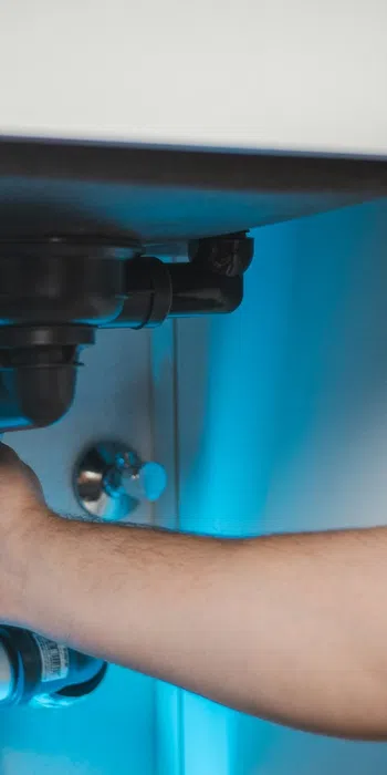 Plumber fixing white PVC pipes under a sink illuminated by blue light in a modern kitchen or bathroom.