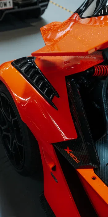 Close-up of an orange sports car rear wing and wheel with carbon fiber details and raindrops.