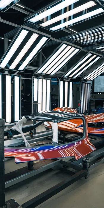 Brightly lit automotive paint booth with a red and blue car body part undergoing finishing touches.