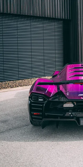 Rear view of a vibrant purple KTM X-Bow sports car parked outside a modern building with black vertical panels.
