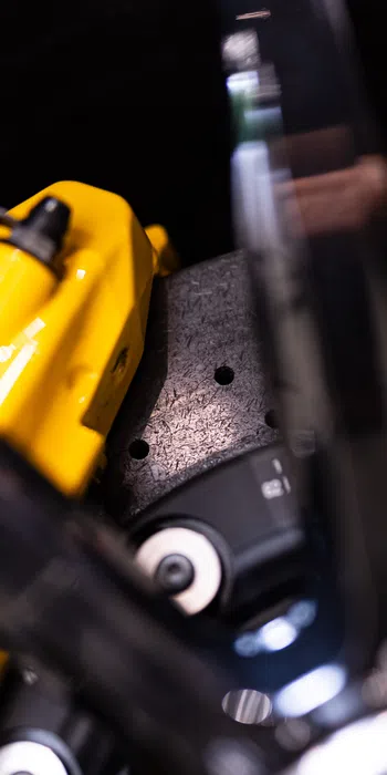 Close-up of a yellow brake caliper, part of a perforated brake disc with a blurred black component in the foreground