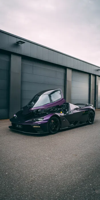 Dark purple sports car with canopy door open parked in front of gray industrial garage doors on overcast day