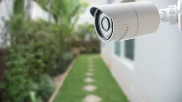 White outdoor security camera mounted on wall overlooking garden path with stepping stones and greenery