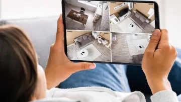 Person holding tablet displaying four different home security camera views inside a modern living space.