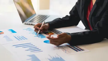 Businessperson analyzing financial charts and graphs with pen and laptop on desk in office.